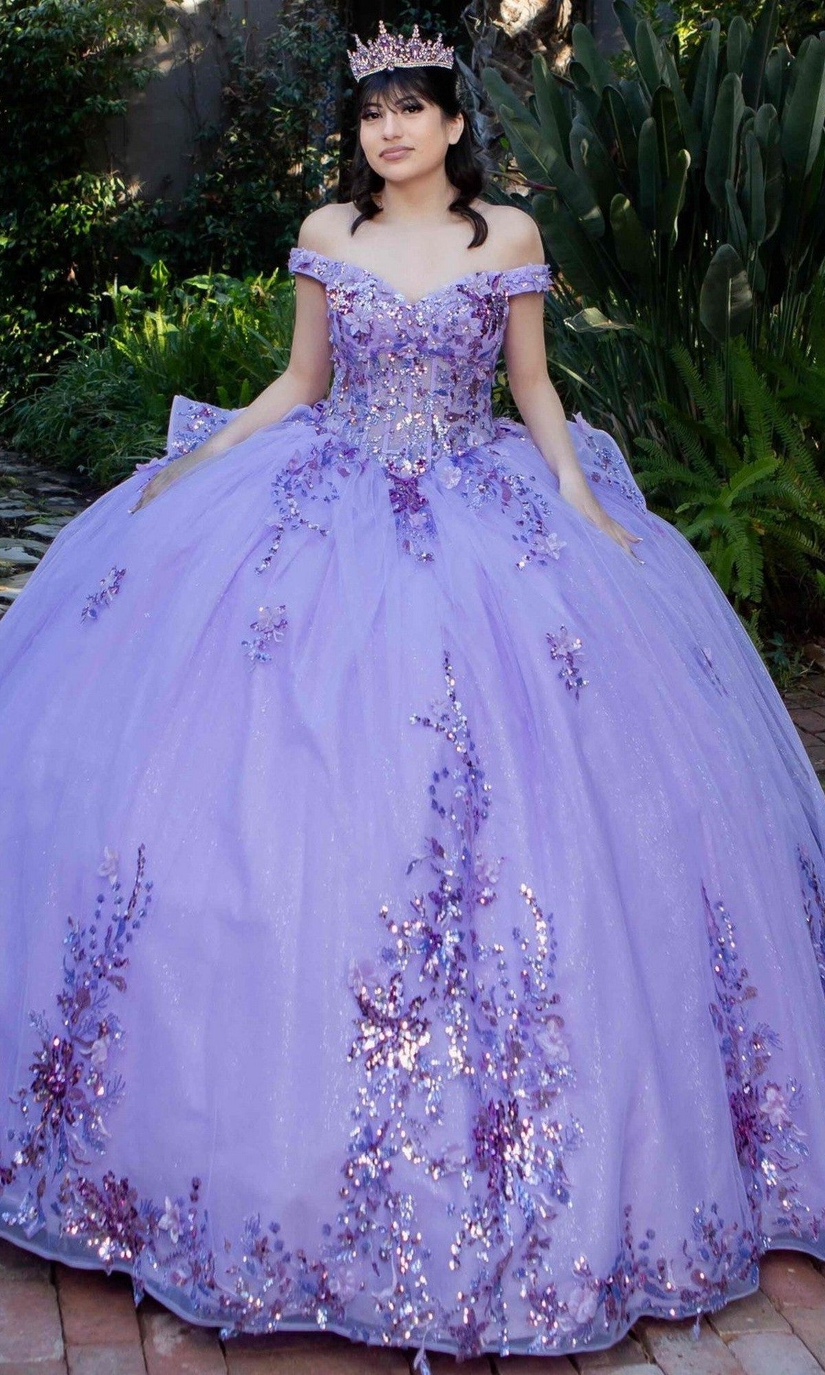A young woman wearing the Quinceanera Ball Gown 8125J, an off-the-shoulder dress with a lace-up corset bodice and sparkling purple details, poses outdoors on a brick path with greenery, complemented by a matching tiara.